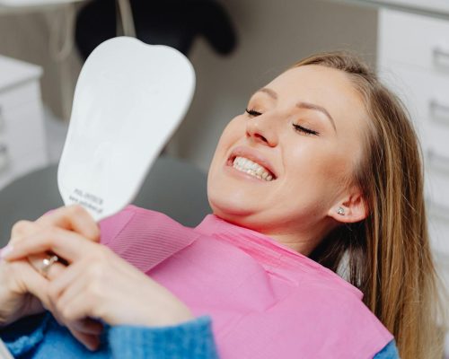 A woman at a dental clinic smiles while holding a mirror, showcasing her dental treatment results.